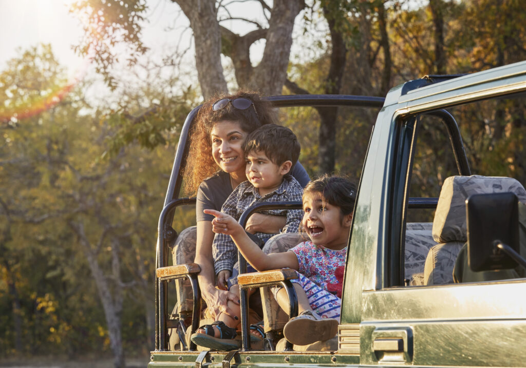 A smiling mother and her two excited young children enjoying a safari ride in an open jeep, with the girl pointing enthusiastically at something in the forest during sunset.